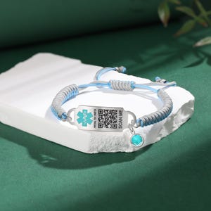 May include: A silver and blue medical ID bracelet with a turquoise charm. The bracelet features a braided design with a metal plate displaying a medical symbol, a QR code, and the text "SCAN ME" and "HEALTH INFO".