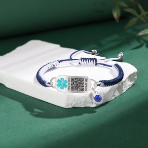 May include: A blue and white braided bracelet with a silver medical ID tag. The tag has a blue medical symbol, a QR code, and the text "HEALTH INFO" and "SCAN ME". A small blue gemstone hangs from the bracelet.