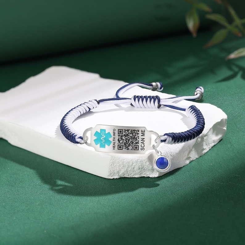 May include: A blue and white braided bracelet with a silver medical ID tag. The tag has a blue medical symbol, a QR code, and the text "HEALTH INFO" and "SCAN ME". A small blue gemstone hangs from the bracelet.