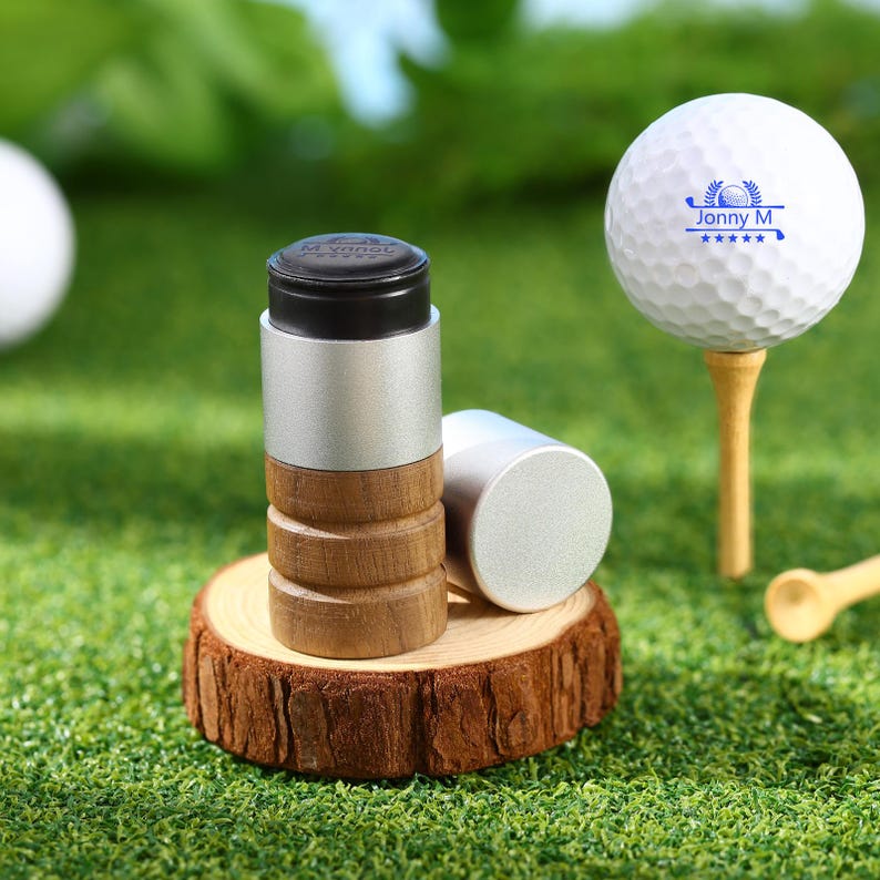 May include: A golf ball on a wooden tee next to a golf ball marker. The marker has a black top, silver body, and wooden base. The golf ball has the text "Jonny M" and a golf club logo.