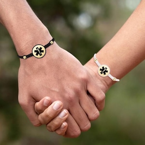 Może przedstawiać: Dwa pasujące bransoletki z czarnymi i białymi sznurkami i złotymi zawieszkami z symbolem medycznym. Bransoletki są noszone na nadgarstkach dwóch osób trzymających się za ręce.