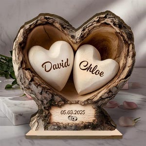 May include: A wooden heart-shaped sculpture with two white heart-shaped carvings inside. The carvings are inscribed with the names "David" and "Chloe". A wooden plaque at the base displays the date "05.03.2025" and two wedding rings.