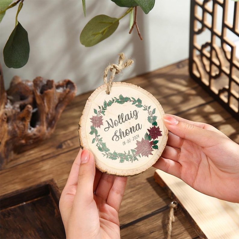 May include: A round wooden ornament with the text "Nollaig Shona" and the date 01-02-2024, surrounded by a green and burgundy floral wreath. The ornament is held by a person. A natural twine loop is attached for hanging.