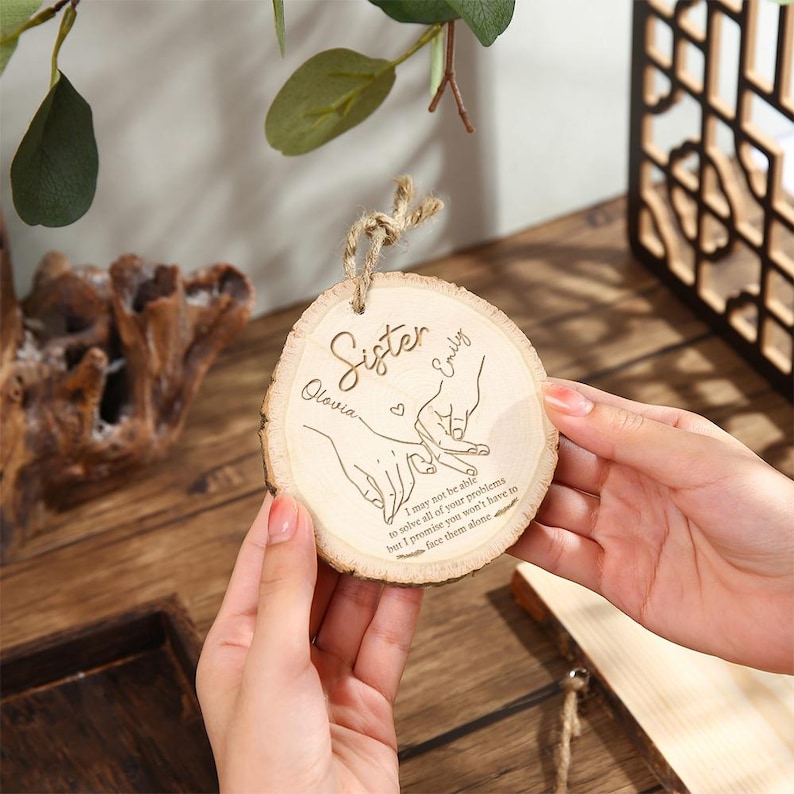 May include: A round wooden ornament with a natural wood grain. The ornament has the word "Sister" and the names "Olovia" and "Emily" engraved on it, along with a drawing of two hands holding each other. The text "I may not be able to solve all of your problems but I promise you won't have to face them alone" is also engraved.