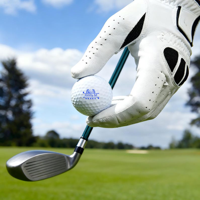 May include: A white golf ball with the text "Jonny M" and a logo is held by a gloved hand above a golf club. The golf club has a silver head and a green shaft. The background shows a green golf course and a blue sky.