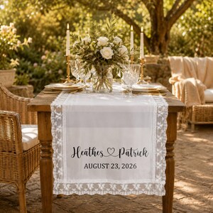 Pode incluir: Um caminho de mesa branco com bordas de renda intrincadas, apresentando o texto impresso em preto "Heathes ♡ Patrick" e "AUGUST 23, 2026" centralizado no tecido. O caminho está colocado sobre uma mesa de madeira com um arranjo floral e velas.