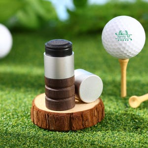 May include: Golf accessories set on a green surface. The set includes a golf ball on a tee, a cylindrical golf ball marker with a black top, silver body, and wooden base, and a separate silver cap. The golf ball has the text "Jonny M" and a logo.
