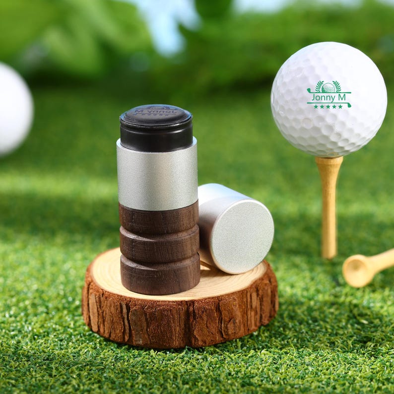 May include: Golf accessories set on a green surface. The set includes a golf ball on a tee, a cylindrical golf ball marker with a black top, silver body, and wooden base, and a separate silver cap. The golf ball has the text "Jonny M" and a logo.
