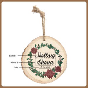 May include: A round wooden ornament with a natural wood grain finish. The ornament features a green wreath with red poinsettias and the text "Nollaig Shona" and the date "01-02-2024". A jute twine loop is attached for hanging.