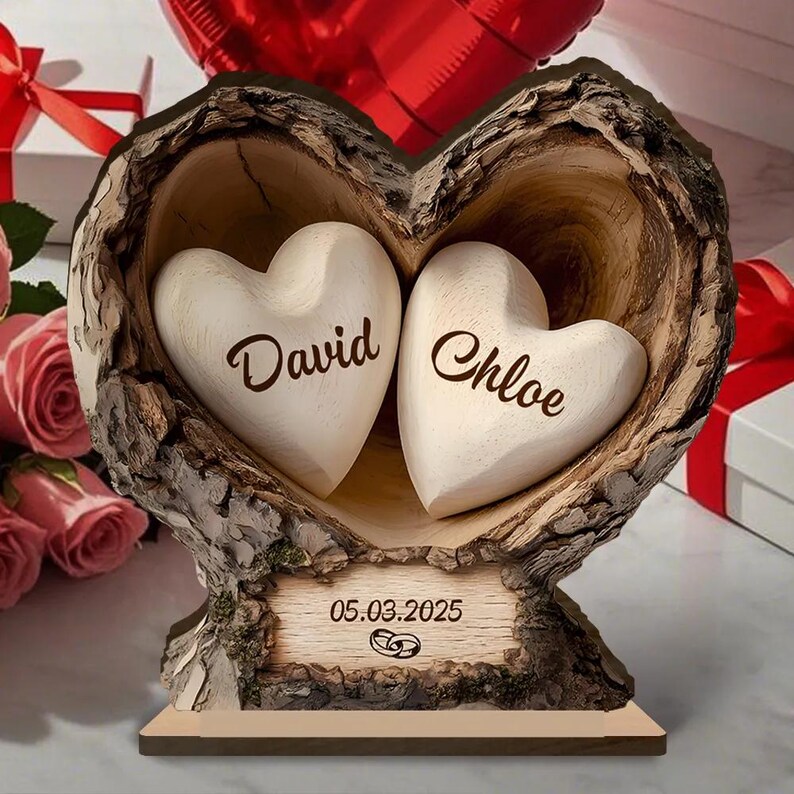 May include: A wooden heart-shaped sculpture with two white hearts inside, each bearing a name. The sculpture is carved from a tree trunk, with a base that includes the date "05.03.2025" and wedding rings. The background includes roses and a red heart balloon.