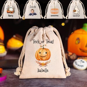 Peut inclure: Sacs en toile beige avec des motifs sur le thème d'Halloween. Un sac présente une citrouille souriante avec le texte "Trick or Treat !" et le nom "Isabelle". D'autres sacs affichent un fantôme, une photo d'enfant, une citrouille et un fantôme avec un seau de citrouille, chacun avec un nom.