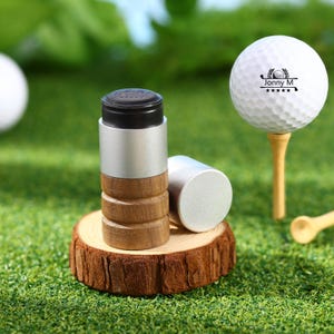 May include: A golf-themed product set featuring a white golf ball on a wooden tee, and two cylindrical golf ball markers. The markers have a silver top and bottom with a wooden midsection. The golf ball has the text "Jonny M" and a golf club logo.