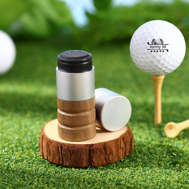 May include: A golf-themed product set featuring a white golf ball on a wooden tee, and two cylindrical golf ball markers. The markers have a silver top and bottom with a wooden midsection. The golf ball has the text "Jonny M" and a golf club logo.