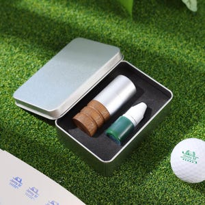 May include: A silver metal box with a black interior holds a golf ball marker and a small bottle of green liquid. A golf ball with the text "Jonny M" is also visible on a green surface.
