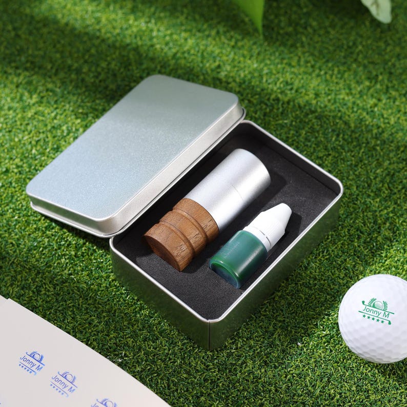 May include: A silver metal box with a black interior holds a golf ball marker and a small bottle of green liquid. A golf ball with the text "Jonny M" is also visible on a green surface.