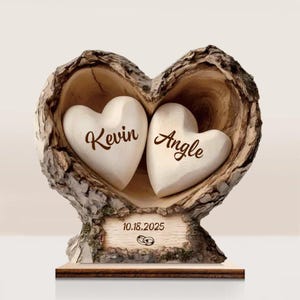 May include: A heart-shaped wooden sculpture with two white heart-shaped stones inside. The names "Kevin" and "Angle" are inscribed on the stones. The date "10.18.2025" and wedding rings are on a wooden plaque at the base.