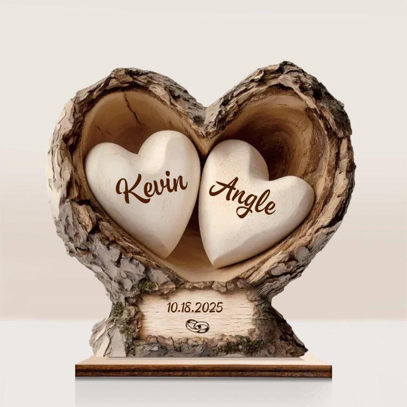 May include: A heart-shaped wooden sculpture with two white heart-shaped stones inside. The names "Kevin" and "Angle" are inscribed on the stones. The date "10.18.2025" and wedding rings are on a wooden plaque at the base.