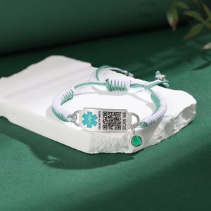 May include: A medical ID bracelet with a white and green braided cord. The silver-toned tag has a medical symbol and a QR code, with the text "HEALTH INFO" and "SCAN ME". A green gemstone charm hangs from the bracelet.