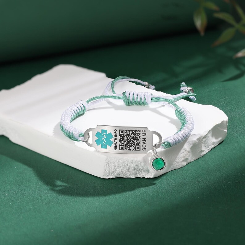 May include: A medical ID bracelet with a white and green braided cord. The silver-toned tag has a medical symbol and a QR code, with the text "HEALTH INFO" and "SCAN ME". A green gemstone charm hangs from the bracelet.