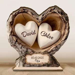 May include: A wooden heart-shaped sculpture with two white hearts inside, each bearing a name in elegant script. The sculpture is carved from a tree trunk, with a base that includes the date 05.03.2025 and wedding rings.