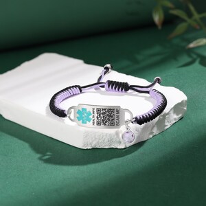 May include: A medical ID bracelet with a braided cord in black and lavender. The silver-tone ID tag has a medical symbol and a QR code. A small, round, purple charm hangs from the bracelet. The text "HEALTH INFO" and "SCAN ME" are visible.