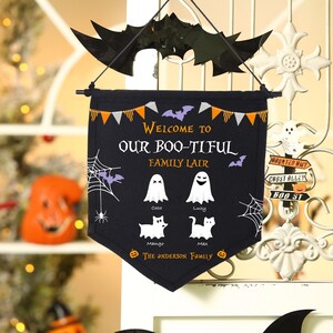 Puede incluir: Bandera negra de Halloween con el texto "WELCOME TO OUR BOO-TIFUL FAMILY LAIR". La bandera presenta fantasmas, murciélagos, arañas y los nombres Coco, Lucy, Mango y Max. Un murciélago negro cuelga encima.
