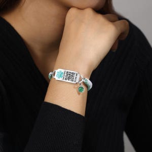 May include: A medical ID bracelet with a white and green woven band. The silver-toned ID plate features a medical symbol and a QR code, with the text "HEALTH INFO" and "SCAN ME". A small green charm hangs from the bracelet.
