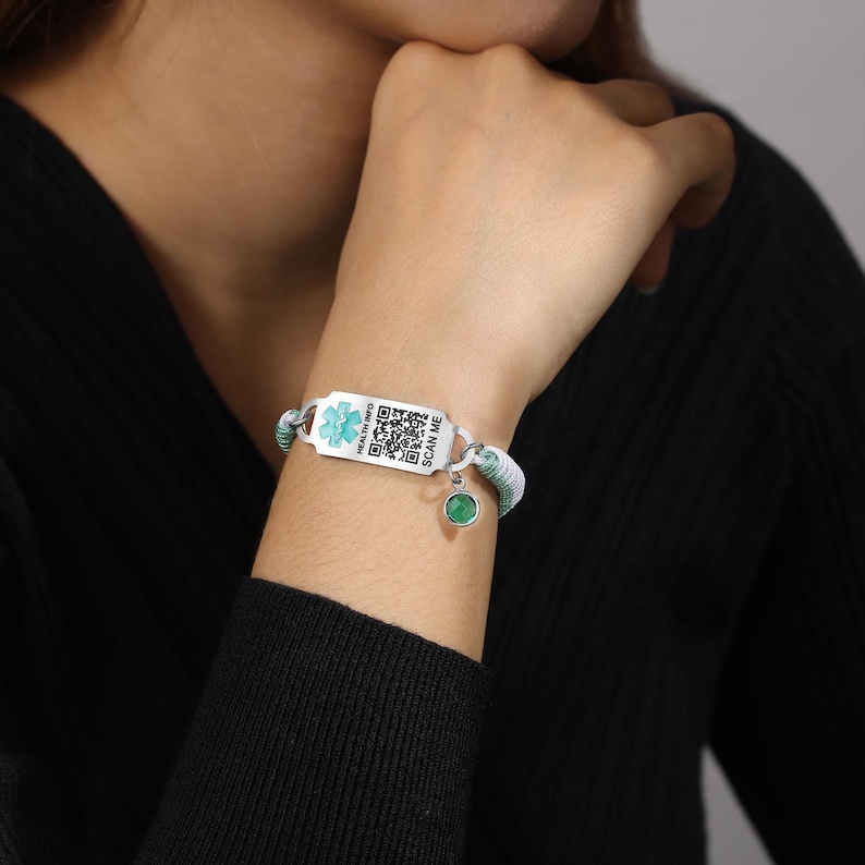 May include: A medical ID bracelet with a white and green woven band. The silver-toned ID plate features a medical symbol and a QR code, with the text "HEALTH INFO" and "SCAN ME". A small green charm hangs from the bracelet.