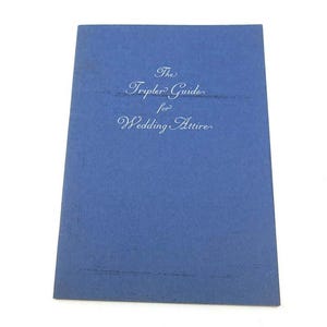 May include: A rectangular blue booklet with the text The Tripler Guide for Wedding Attire printed in white script font on the front cover. The cover has a textured surface and the title is centered in the upper portion of the blue booklet.