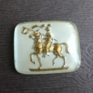 May include: A rectangular, vintage-style brooch with a gold-colored equestrian design. The rider and horse are detailed against a translucent, off-white background. The brooch has a slightly rounded shape.