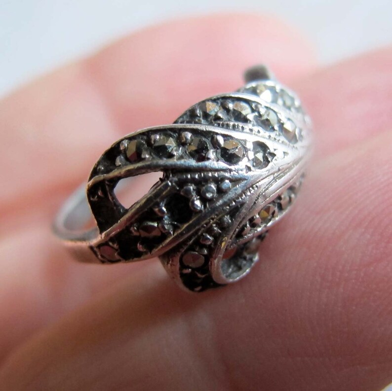 Puede incluir: Un anillo de tono plateado con un dise&ntilde;o de remolinos en forma de hoja, incrustado con numerosas piedras de marcasita peque&ntilde;as, oscuras y facetadas. La banda de metal tiene un acabado pulido y la secci&oacute;n superior decorativa muestra un patr&oacute;n texturizado.