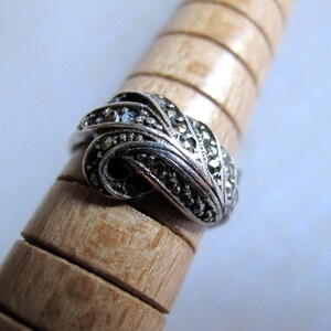 Puede incluir: Un anillo de plata con un dise&ntilde;o trenzado en forma de nudo, incrustado con peque&ntilde;as piedras de marcasita oscuras y facetadas. La banda tiene un acabado met&aacute;lico oxidado y se muestra sobre un mandril de madera con ranuras horizontales.