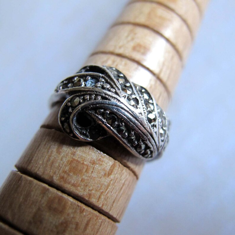 Puede incluir: Un anillo de plata con un dise&ntilde;o trenzado en forma de nudo, incrustado con peque&ntilde;as piedras de marcasita oscuras y facetadas. La banda tiene un acabado met&aacute;lico oxidado y se muestra sobre un mandril de madera con ranuras horizontales.