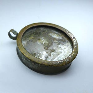 May include: A round, brass-toned metal pendant featuring a weathered, tarnished finish and a small circular loop on one side. The center contains a clear, slightly cloudy glass insert with visible signs of wear, scratches, and internal residue.