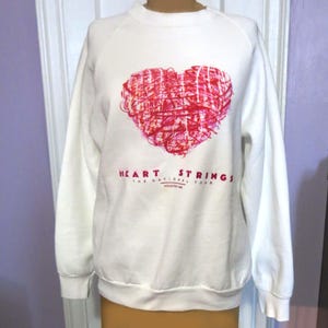 May include: White crew neck sweatshirt featuring a large red heart design composed of scribbled lines. The text "HEART STRINGS THE NATIONAL TOUR HOUSTON 1988" is printed below the heart in red. The sweatshirt has long sleeves.