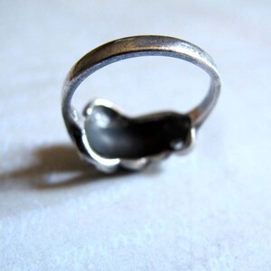 Puede incluir: Un anillo de metal plateado con una banda lisa y redondeada y una secci&oacute;n superior curva de forma org&aacute;nica. La superficie met&aacute;lica muestra una p&aacute;tina oscura y ligeramente desgastada con reflejos sutiles a lo largo de los bordes de la banda.