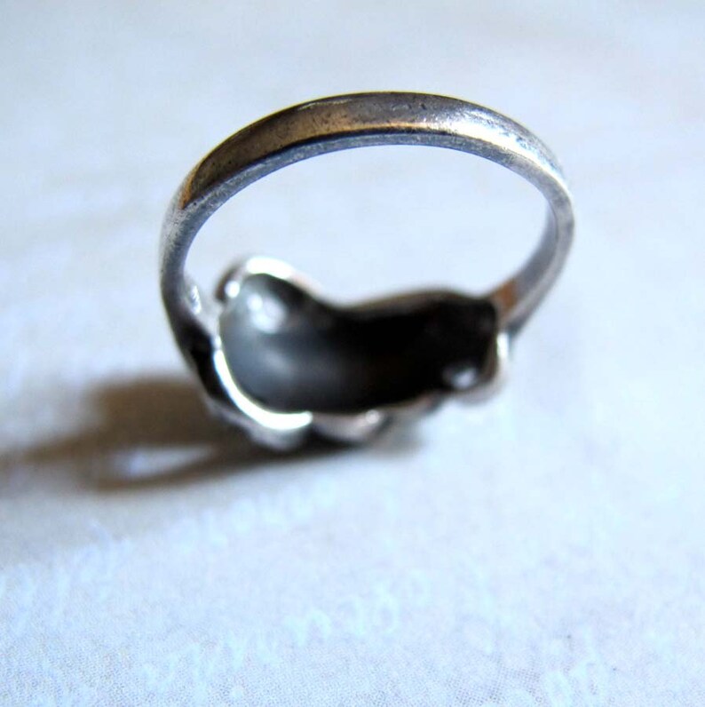 Puede incluir: Un anillo de metal plateado con una banda lisa y redondeada y una secci&oacute;n superior curva de forma org&aacute;nica. La superficie met&aacute;lica muestra una p&aacute;tina oscura y ligeramente desgastada con reflejos sutiles a lo largo de los bordes de la banda.
