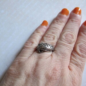 Puede incluir: Un anillo de tono plateado con un dise&ntilde;o de remolinos superpuestos, incrustado con numerosas piedras peque&ntilde;as, oscuras y facetadas. El metal tiene un acabado oxidado que resalta las curvas intrincadas y la superficie texturizada del aro.