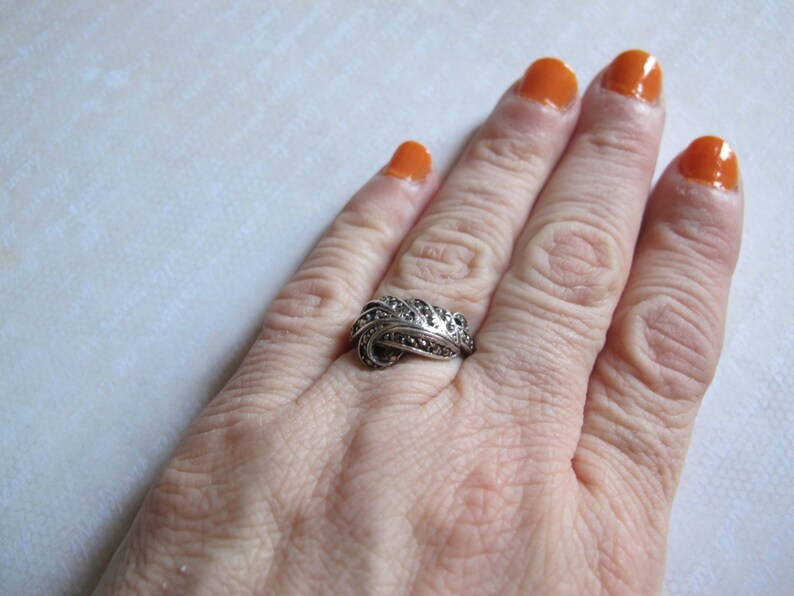 Puede incluir: Un anillo de tono plateado con un dise&ntilde;o de remolinos superpuestos, incrustado con numerosas piedras peque&ntilde;as, oscuras y facetadas. El metal tiene un acabado oxidado que resalta las curvas intrincadas y la superficie texturizada del aro.