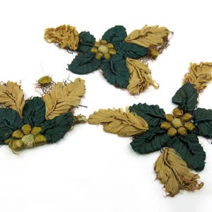 May include: Three floral embellishments with dark green leaves and tan petals. The center of each flower has small, round, yellow-green accents. The embellishments are arranged on a white background.