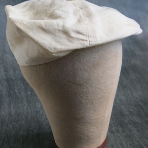 May include: A light beige flat cap made of a woven fabric, featuring a rounded crown, a short stiff brim at the front, and a small fabric-covered button at the center top. The cap has visible stitched seams connecting the panels.
