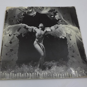 May include: Black and white photograph of a person with feathered wings, arms outstretched. The person is standing in front of a dark background, with a row of candles at the bottom. The image is on a square, aged paper.