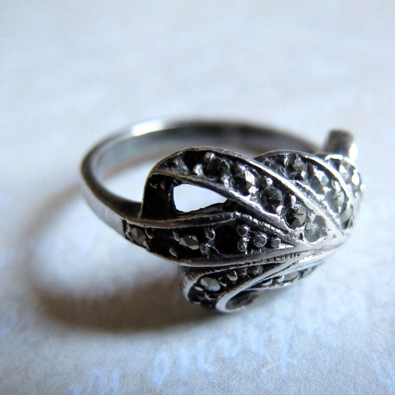 Puede incluir: Un anillo de tono plateado que presenta un elemento de dise&ntilde;o curvo en forma de hoja en la parte frontal. La superficie del adorno met&aacute;lico est&aacute; incrustada con numerosas piedras peque&ntilde;as, oscuras y facetadas que crean un aspecto texturizado.