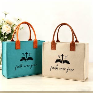May include: Two tote bags, one teal and one beige, with brown handles and orange accents. Each bag features a black graphic of a cross, open book, and the words "faith over fear."