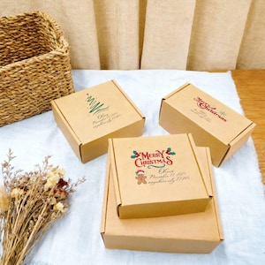 Puede incluir: Cuatro cajas pequeñas y cuadradas de cartón marrón con diseños navideños. Una caja presenta un árbol de Navidad verde, otra tiene "Merry Christmas" en rojo y la tercera un hombre de jengibre. Las cajas están dispuestas sobre una superficie blanca.