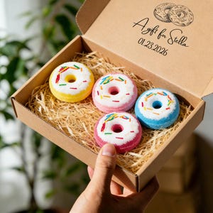 May include: A cardboard gift box holding four donut-shaped bath bombs in yellow, pink, and blue, with white frosting and colourful sprinkles. The box is filled with straw and has the text "A gift for Stella 01.23.2026".