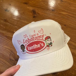 May include: White baseball cap with the text "I got a Lobotomy at Ianthe's" in red and black. The cap features cartoon illustrations and hearts. The cap has a curved brim and a mesh back.