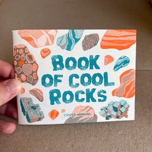Peut inclure: Un livret rectangulaire intitulé Book of Cool Rocks par Casper Manning. La couverture blanche présente un motif répétitif de roches illustrées dans des tons orange, bleu sarcelle et marron, avec le titre en grandes lettres texturées.