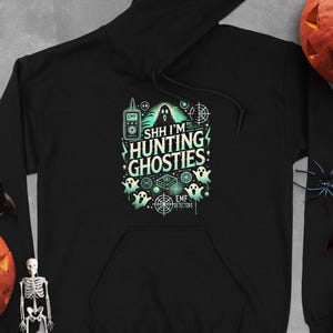 Könnte beinhalten: Schwarzer Hoodie mit einem grün-weißen Grafikdruck von Geistern, EMF-Detektoren und dem Text "Shh I'm Hunting Ghosties".
