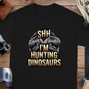 Puede incluir: Camiseta negra con un gráfico de dos dinosaurios y el texto "Shh I'm Hunting Dinosaurs".